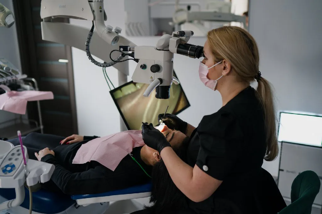 Dr. Ramona Drilea, în costum medical negru, lucrând la microscop cu o pacientă întinsă în fotoliul stomatologic